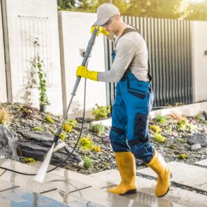 Caucasian Men in Waterproof Yellow Gumboots and Gloves Cleaning the Concrete Terrace around the House with Pressure Washer. Property Care and Maintenance Theme.